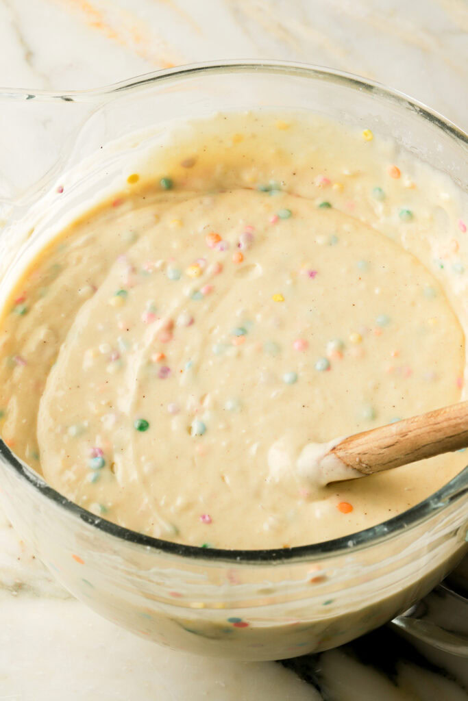 vanilla cake batter with sprinkles mixed into it, in glass mixing bowl with spatula