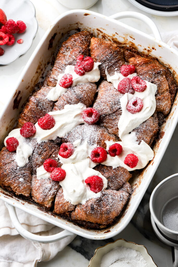 baked croissant french toast in casserole dish and topped with whipped cream and raspberries