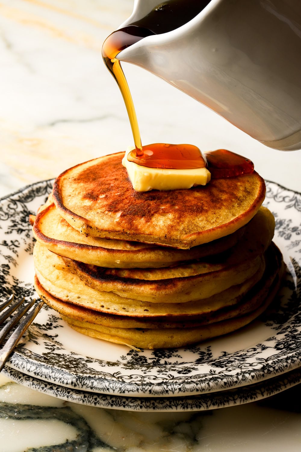 stack of pancakes on a black and white plate with butter on top and maple syrup pouring out of a spout
