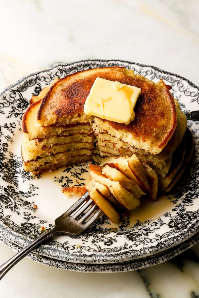 stack of pancakes on a black and white plate with a slice taken out and butter and maple syrup on top