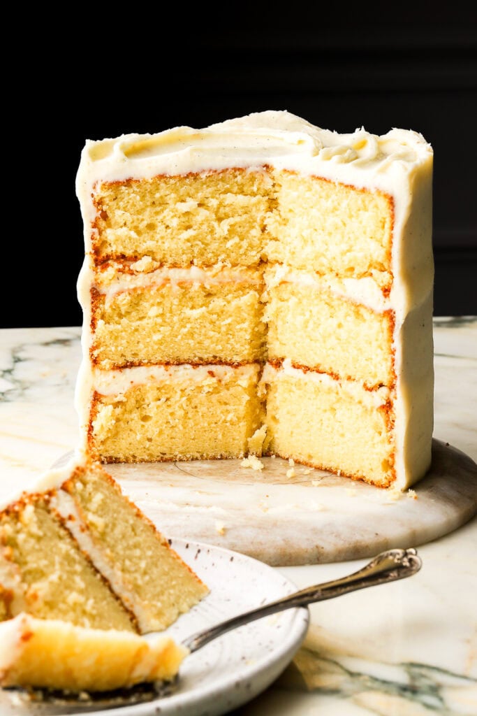 three layer vanilla cake with vanilla frosting sliced showing the inside with a black background a plate with a slice in front of it