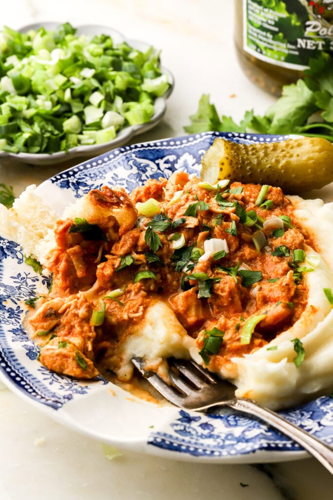 blue and white plate with a fork going through mashed potatoes with chicken stew (tocanita) on top. Topped with parsley, chives and a bite out of a pickle on the side
