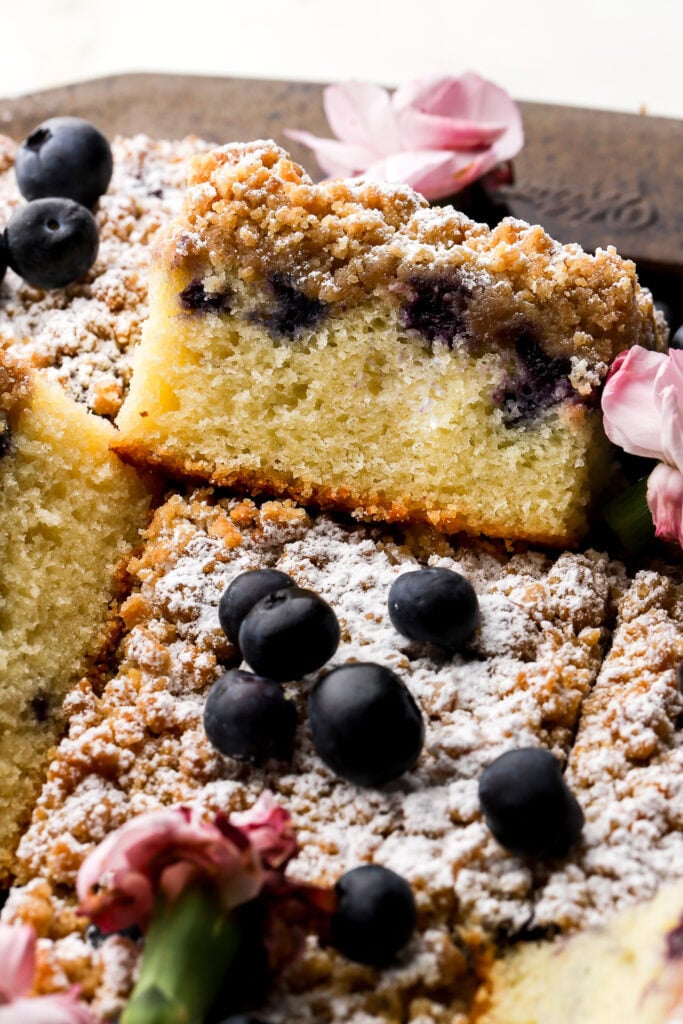 close up of a slice of blueberry coffee cake sitting up halfway on another one topped with blueberries