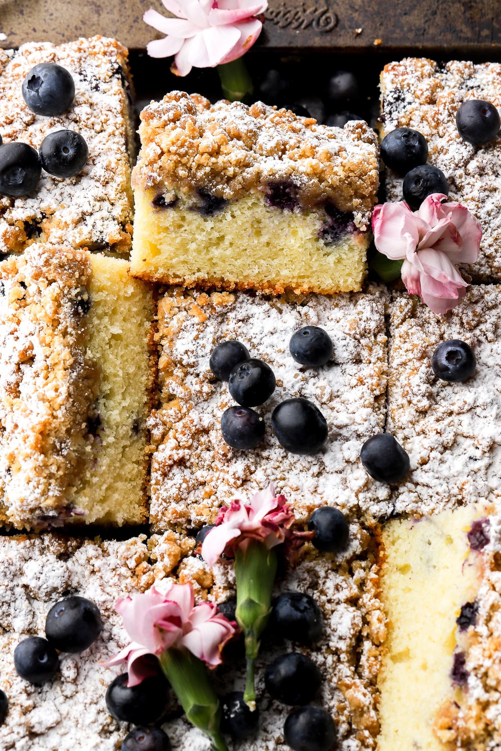 blueberry coffee cake with brown sugar streusel dusted with powdered sugar and sliced with some one top of each other - blueberries and pink flowers thrown around
