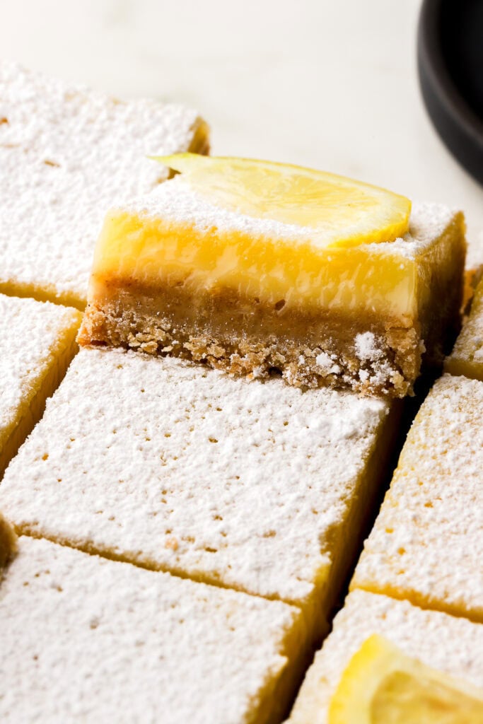 lemon bars dusted with powdered sugar and sliced, one sitting up higher on top of the others ones, with a lemon slice on top, to show the inside texture