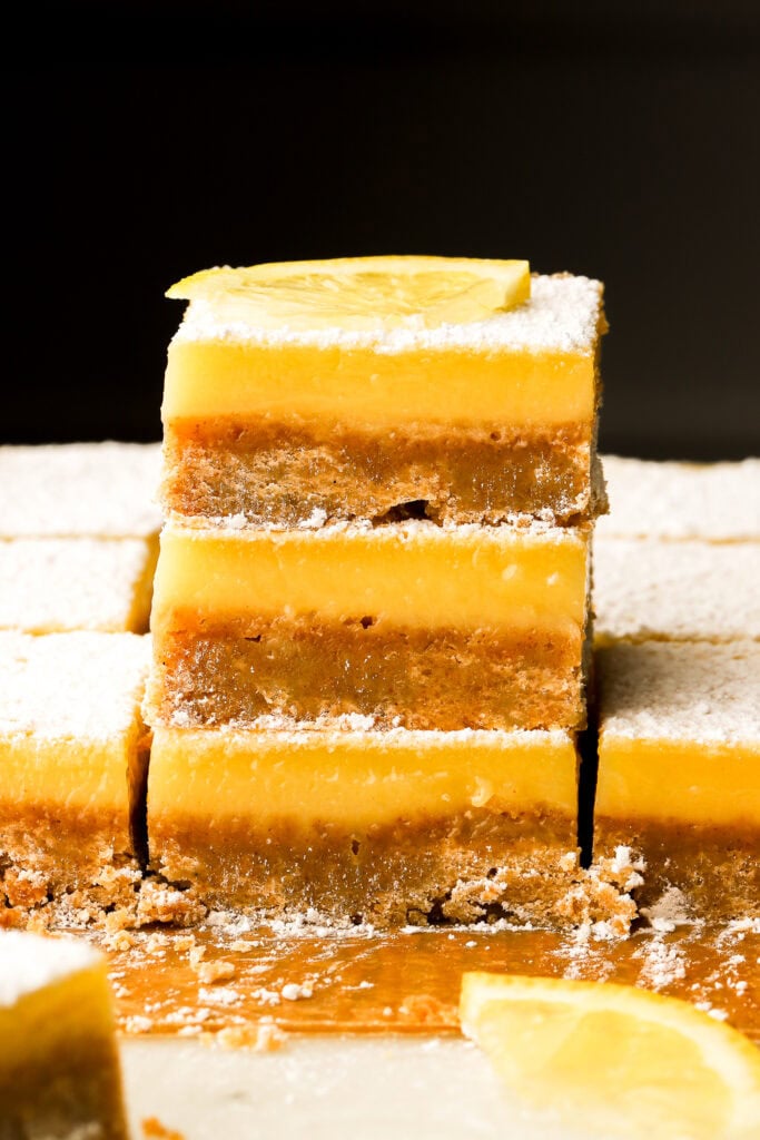 three lemon bars sliced and stacked on top of each other with a slice of lemon on top and a black background