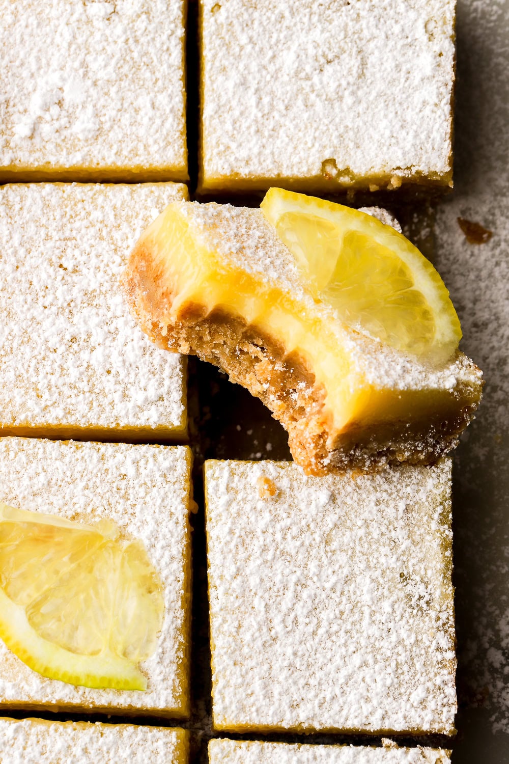 overhead shot of lemon bars, dusted with powdered sugar and sliced - two with lemon slices on top and one with a bite taken out of it, facing up