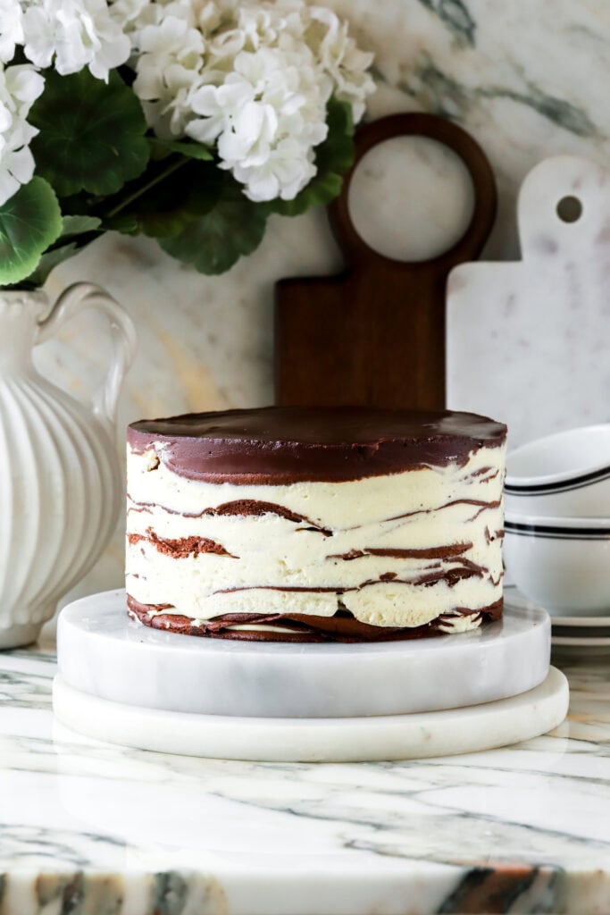 finished reverse dobos cake with 16 layers, topped with chocolate ganache - on a marble countertop and florals and cutting boards in the background. 