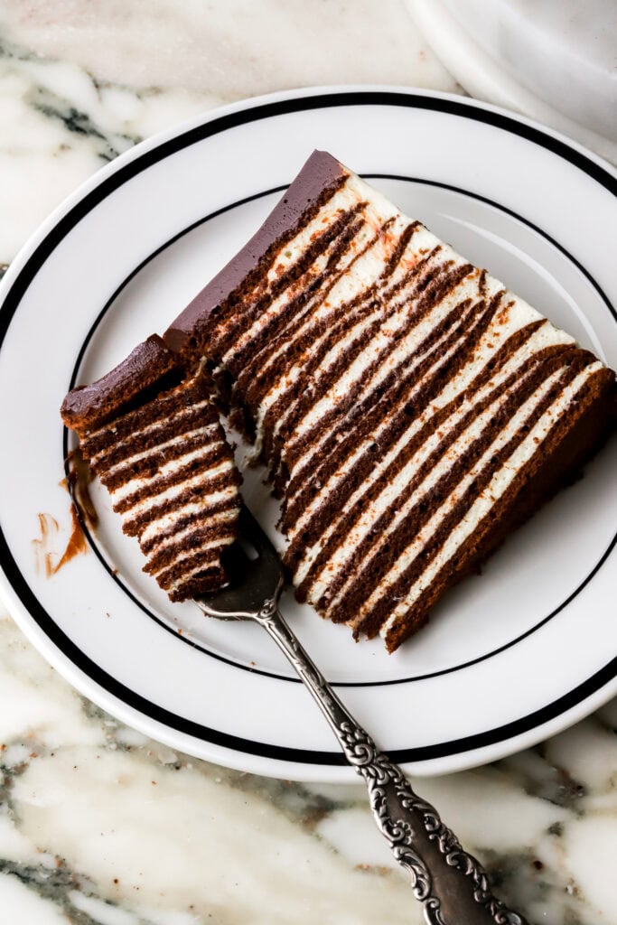 close up of slice of cake on a black and white plate and marble countertop with 16 chocolate cake layers and vanilla cream with a fork taking a bite out. 