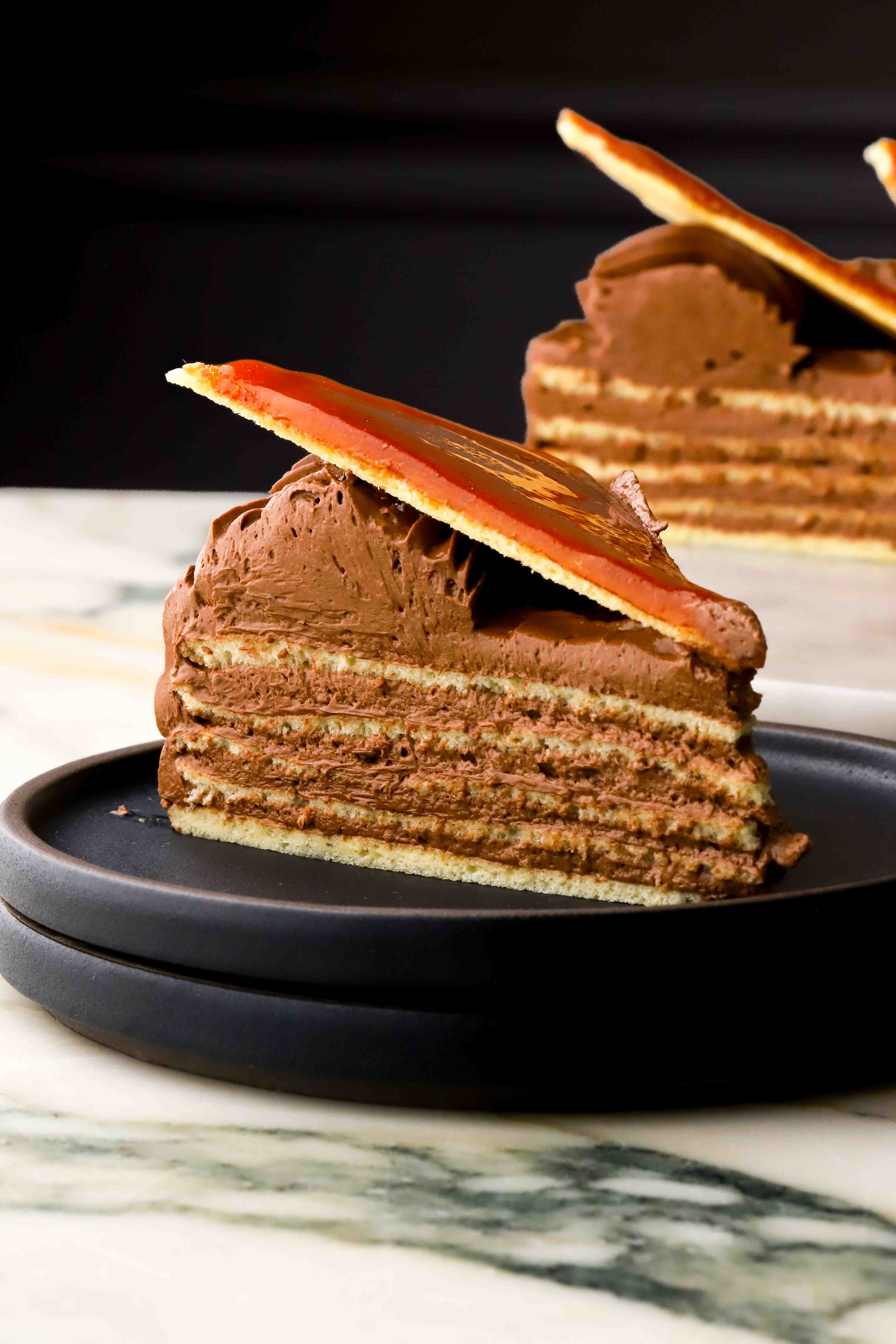 close up of a slice of authentic dobos on a plate with 5 layers of sponge, chocolate buttercream and a caramelized sugar triangle on top
